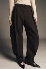 Pantalón cargo de algodón color marrón oscuro, tiro medio, con pliegues frontales, bolsillos delanteros ocultos y bolsillos de plastrón con solapa y botones a presión en las piernas. El bajo del pantalón tiene una trabilla con botón y se muestra doblado hacia afuera.