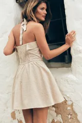 Vestido corto a cuadros beige y blanco, con cuello halter que se ata en la nuca, escote recto con abertura y falda acampanada. Tiene bolsillos laterales ocultos, espalda elastizada y botones decorativos en el frente.