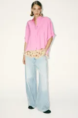 Camisa rosa de cuello solapa y manga corta con vuelta. Bajo asimétrico con aberturas laterales. Cierre frontal con botones ocultos por solapa.