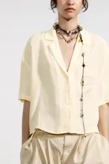 Camisa color amarillo pálido de tejido fluido, con cuello solapa, escote pico, manga corta, bolsillo de parche en el pecho y cierre frontal con botones.