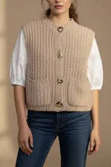 Chaleco color beige de tejido de punto grueso con cuello redondo y cierre frontal con botones dorados. Tiene dos bolsillos frontales de parche.