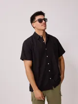 Camisa negra de manga corta con cuello clásico y cierre frontal con botones blancos. Corte recto y holgado.