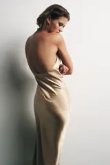 Vestido midi color dorado, con cuello halter ajustable con botón y hombros descubiertos. Detalle de tejido efecto arrugado, espalda pronunciada, bajo asimétrico y cierre lateral con cremallera oculta.