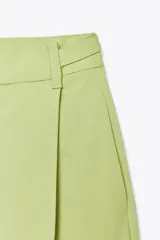 Pantalón verde claro de tiro alto con trabillas y botón en la cintura. Tiene pliegues, bolsillos laterales y falsos bolsillos de vivo en la espalda. El cierre frontal es con cremallera, botón interior y ganchos metálicos.