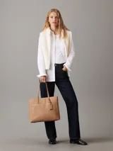 Cartera tote Calvin Klein color tostado, de tamaño mediano, confeccionada en cuero sintético granulado con textura de logo monograma en el bolsillo delantero. Tiene asas fijas, cierre magnético, bolsillo exterior, compartimento interior con cremallera y colgante extraíble con logo.