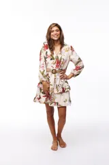 Vestido corto blanco con estampado floral en tonos rojos, verdes y amarillos. Tiene escote en V, mangas largas abullonadas y falda con volados. Se ajusta a la cintura con un cinturón de soga con hebilla circular.
