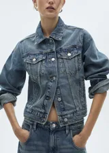 Chaqueta de jean azul de corte recto con cuello camisero, cierre frontal con botones metálicos, dos bolsillos con solapa en el pecho y mangas largas con puños abotonados.