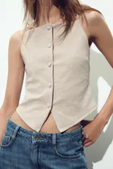 Top tipo chaleco color crema con escote recto y tirantes finos. Tiene cierre frontal con botones a tono y bajo asimétrico con abertura.