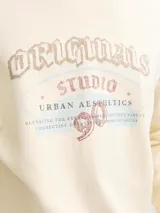 Buzo de manga larga color blanco antiguo (off-white) con cuello redondo y fit holgado. Presenta un estampado frontal con texto que incluye "ORIGINALS STUDIO", "URBAN AESTHETICS" y el número "90" en tonos rosados y dorados.
