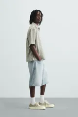 Bermuda de denim de algodón, color gris claro lavado, de corte ancho (baggy fit) y largo hasta la rodilla. Presenta cinco bolsillos y cierre frontal con cremallera y botón.