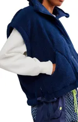 Chaleco de piel sintética tipo sherpa en color blanco crema, con cuello alto y cierre frontal mediante botones a presión. Presenta un diseño de hombros caídos y detalles de lazos decorativos en los laterales inferiores.