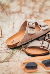 Sandalias de cuero sintético color beige con doble tira y hebillas metálicas ajustables.