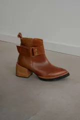 Bota corta de cuero marrón con puntera redondeada y taco cuadrado. Presenta una correa con hebilla alrededor del tobillo y cierre lateral.