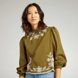 Blusa color verde oliva con cuello mao, mangas largas abullonadas con puños elásticos y bordados florales blancos en el frente.