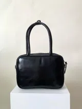 Cartera rígida negra de cuero con tachas plateadas y tres combinaciones de cuero diferentes que aportan textura. Tiene herrajes y cierre metálico plateado marca YKK. Cuenta con un asa fina para llevar al hombro o en la mano y una correa larga extra regulable.