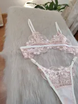 Conjunto de lencería de dos piezas, compuesto por un corpiño tipo bralette y una bombacha tipo colaless, ambos confeccionados en encaje floral color coral/rosado pálido con detalles y elásticos en blanco.