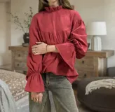 Blusa color beige de corte holgado, cuello alto con volados y mangas largas abullonadas con elástico en los puños.