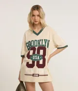 Remera oversize color crema con cuello en V y mangas cortas con ribete verde. Estampado frontal con la palabra "Brooklyn" en verde, el número "98" en bordó con la inscripción "USA" en blanco, y la frase "New York 1998" en blanco.