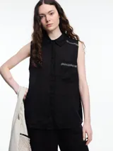 Camisa sin mangas negra de lino con cuello clásico, cierre frontal abotonado y bolsillo de parche en el pecho. Presenta bordados tipo pespunte en contraste en el frente y canesú trasero.