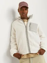 Campera de teddy color blanco hueso, de manga larga y cuello alto. Tiene cierre con cremallera, bolsillos frontales y un bolsillo gris en el pecho con cierre.