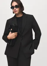 Blazer negro de diseño entallado, con solapas de muesca, hombros estructurados y cierre frontal de un solo botón. Presenta dos bolsillos de ribete delanteros y un bolsillo de ribete en el pecho.