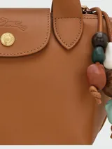 Bolso Longchamp modelo Le Pliage Ceramique XS, de cuero color caramelo, con doble asa corta y correa larga ajustable. Incluye charm colgante de cuentas de madera y cierre con broche a presión.