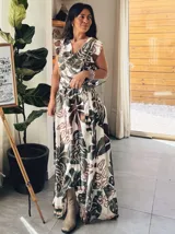Vestido largo con estampado de hojas tropicales en tonos verde y marrón sobre fondo blanco. Tiene escote en V, mangas cortas con volados y corte imperio con lazo ajustable en la cintura.