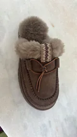 Slippers UGG de cuero color marrón con interior de piel sintética blanca, plataforma color marrón y cordones decorativos.