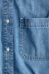 Camisa corta de denim celeste, con cuello americano, botonadura frontal, mangas largas con puños abotonados, bolsillo de parche en el pecho y bajo recto.