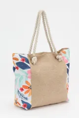 Bolso de playa tipo tote con cuerpo central de tejido rústico color beige (arpillera/yute) y paneles laterales con estampado floral multicolor sobre fondo blanco. Posee asas de cuerda gruesa trenzada.