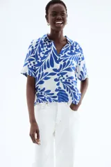 Camisa de manga corta con estampado de hojas y flores en tonos azules sobre fondo blanco. Tiene cuello clásico, botones frontales, corte holgado y canesú con pliegue en la espalda.
