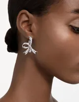 Par de aros colgantes Swarovski con forma de lazo, bañados en rodio y adornados con cristales transparentes engastados. Cierre tipo gancho.