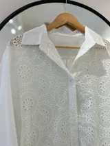 Camisa blanca de manga larga con cuello camisero y cierre frontal con botones. Presenta bordados perforados combinados a tono. El bajo tiene aberturas laterales y la parte trasera es más larga que la delantera.
