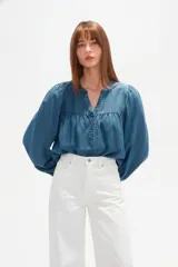 Blusa de lyocell en tono azul denim, con cuello en V, cierre frontal parcial con botones y mangas largas abullonadas con puños elásticos. Presenta un diseño holgado con detalle de fruncido en el pecho.