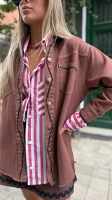 Camisa de denim color chocolate con botones dorados, corte recto, cuello clásico y mangas largas con puños abotonados. Presenta detalles de costuras en contraste y bolsillos frontales.