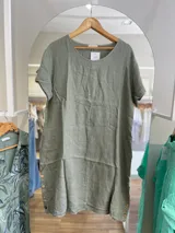 Vestido de lino color verde oliva, corte recto, con cuello redondo y mangas cortas con dobladillo. Presenta botones de madera a lo largo de los laterales.