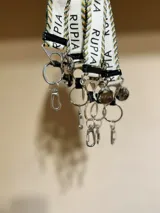 Llavero con cinta de tela estampada con la palabra 'Rupia' en color negro, con detalles en verde y amarillo. Incluye gancho metálico y aro para llaves.