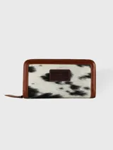 Billetera de cuero con estampado animal print de vaca en blanco y negro, cierre perimetral con cremallera y detalles en cuero marrón.