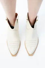 Botas de caña baja color crema, estilo texanas, con bordados a tono en la puntera y caña. Cuentan con un tacón bajo y punta alargada redondeada con suela cuadrada de terminación lisa que sobresale levemente.