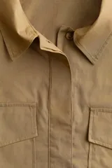Camisa color marrón claro de algodón, corte cropped, con cuello clásico, cierre frontal con botones ocultos, mangas largas con puños abotonados, dos bolsillos con solapa y botón en el pecho y canesú con pliegue en la espalda.