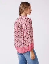 Camisa de gasa con cuello con solapa y apertura central con botones. Presenta un estampado de patrones geométricos en tonos rosados, marrones y crema, con un borde inferior en un patrón geométrico más denso en tonos rojizos/rosados. Las mangas son amplias de largo tres cuartos con puño elastizado.