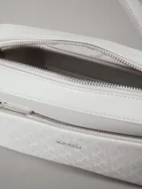Cartera tipo camera bag de color blanco/gris claro, confeccionada en piel sintética con textura de monograma en el bolsillo frontal. Cuenta con dos correas desmontables: una ancha y otra fina, ambas ajustables. El bolsillo frontal tiene el logo de Calvin Klein en metal plateado.