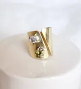 Anillo dorado con diseño abierto y dos piedras incrustadas, una blanca y otra verde.