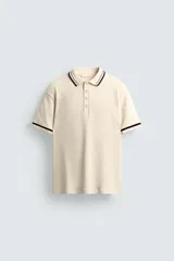 Polo de hombre color crema, de corte regular, con cuello solapa y cierre frontal de botones. Tiene manga corta con rib a contraste en color negro.