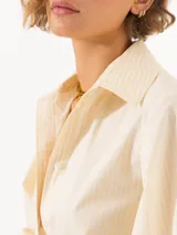 Camisa amarilla confeccionada en poplin rayado con hilos de lurex, mangas abullonadas, botones visibles forrados a tono de tela y detalles de remache Lipstick en la punta del cuello.