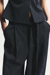 Pantalón negro con finas rayas verticales blancas, de tiro alto y corte ancho. Incluye cinturón de lazo en el mismo tejido, bolsillos laterales y pinzas.