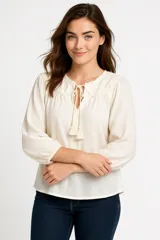 Camisa color crema con cuello a la base, mangas largas abullonadas con puños elásticos, escote en V con cordones y borlas.