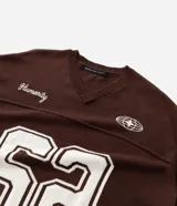 Remera oversized deportiva color marrón con cuello en V, rayas blancas en las mangas y en la parte inferior, y el número "62" estampado en blanco en el frente. Incluye el bordado "Humanity" y un logo de globo terráqueo.