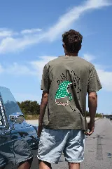 Remera color verde militar con estampado en la espalda de un perro caniche con pañuelo verde con lunares blancos, anteojos de sol y collar de perlas. El estampado incluye la palabra "Bonjour".