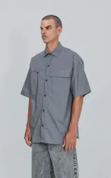 Camisa gris de manga corta confeccionada en poplin 100% algodón, con silueta oversize y dos bolsillos delanteros, uno con solapa y otro con ojal y botón.